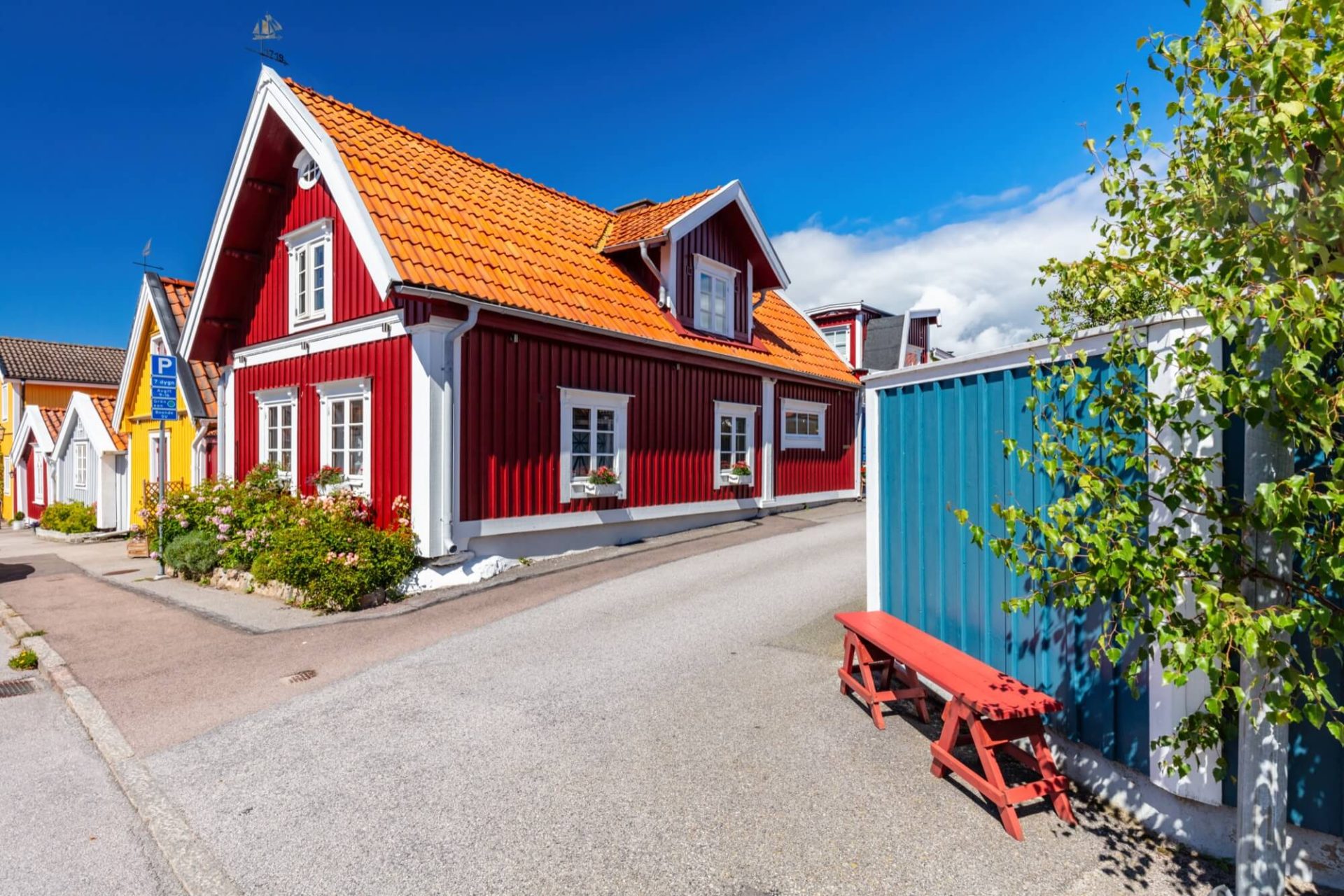 Ett traditionellt rött trähus prytt med vita hörndekorationer och ett orange tak av tegel, beläget i en charmig skärgårdsmiljö